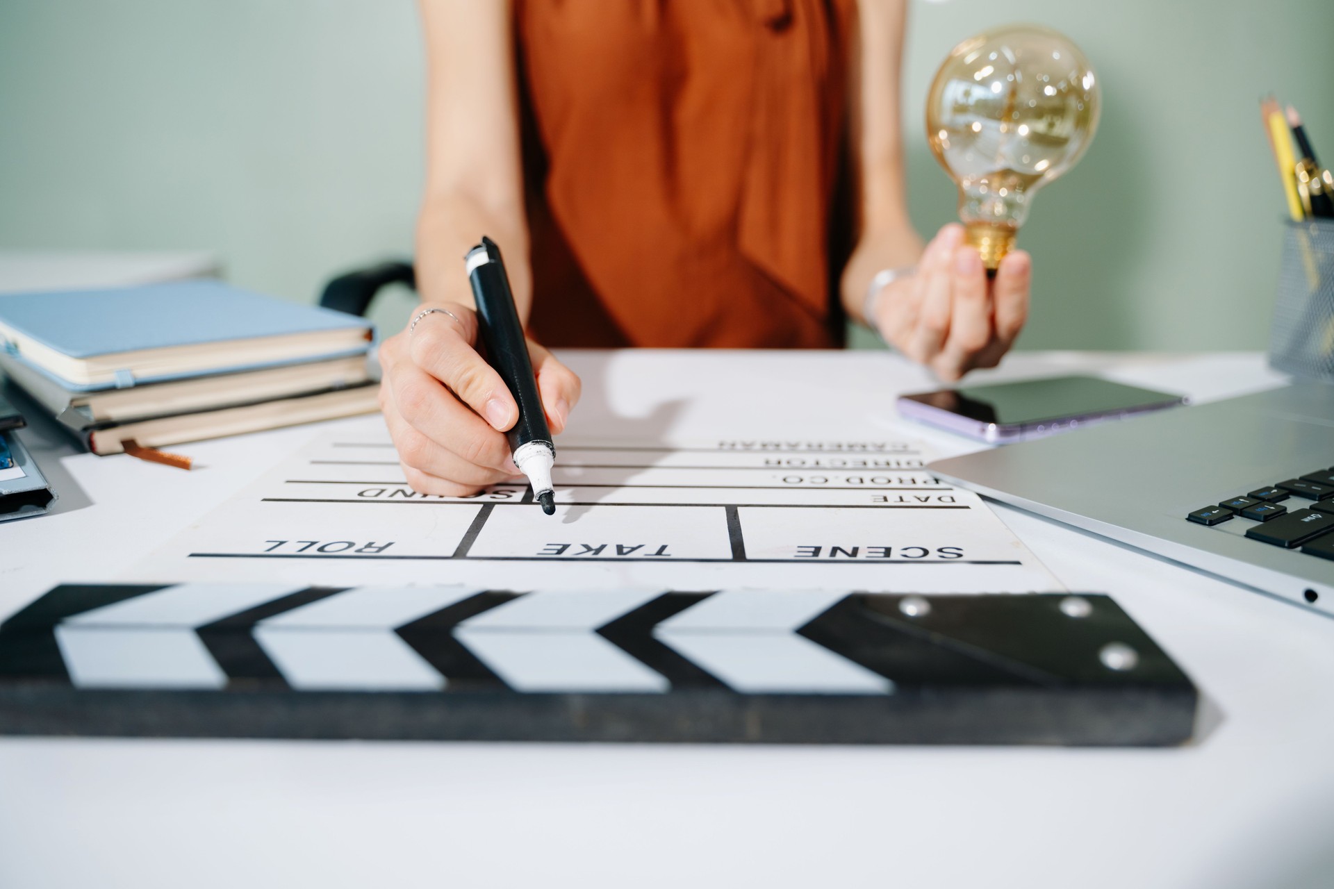 Creative woman holds a glowing lightbulb while using a laptop and clapperboard in modern office. Symbol of inspiration, film production, and innovative ideas.